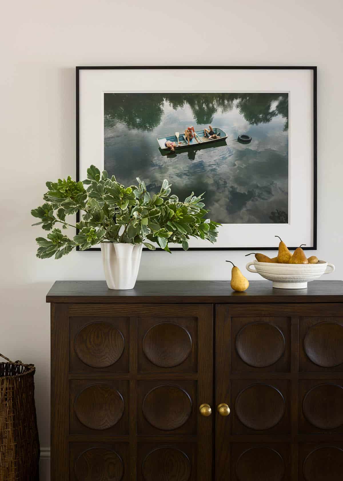 Framed lake photograph above dark wood cabinet styled with pears and greenery