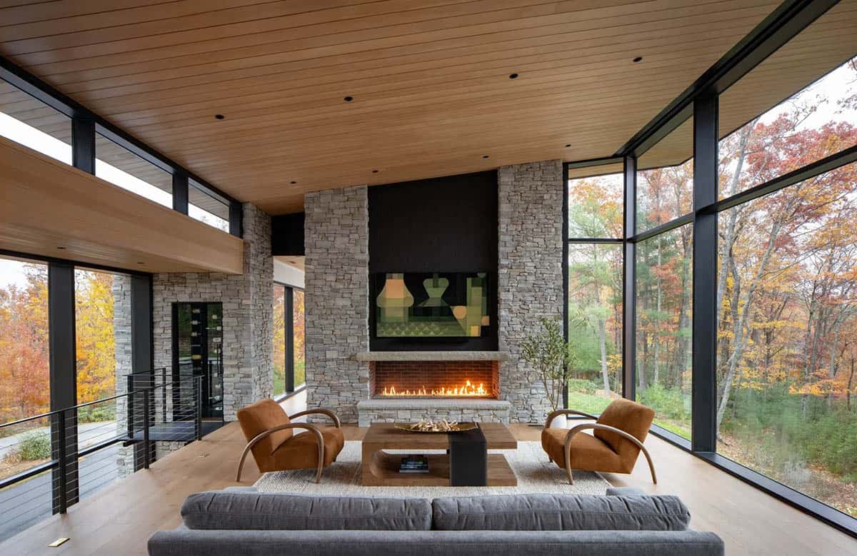 Living room with stone fireplace, wood ceiling, and floor-to-ceiling windows overlooking fall foliage
