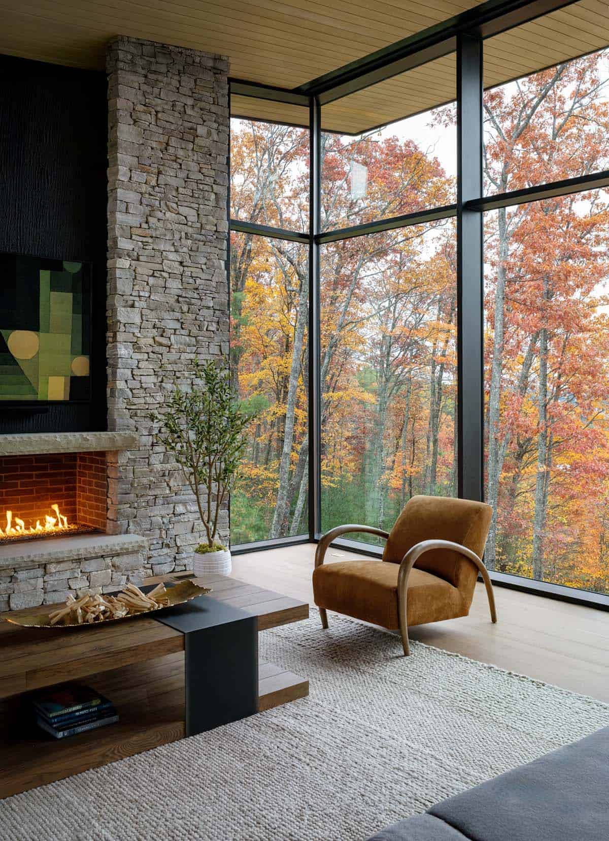 Corner window seating area with stacked stone fireplace and leather armchair, autumn forest view