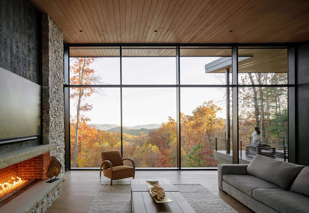 Living room with wraparound windows showing mountain and forest views at dusk