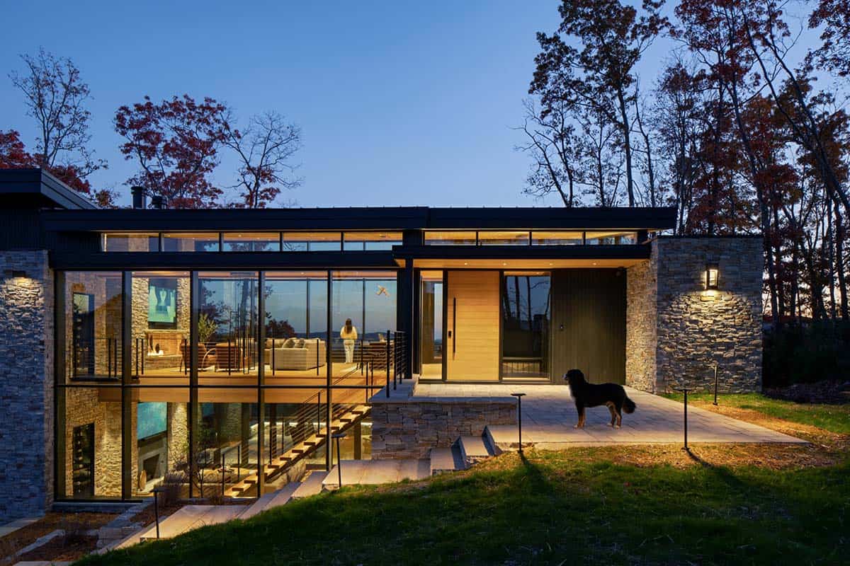 Two-story glass-walled home exterior lit at dusk with dog on stone patio