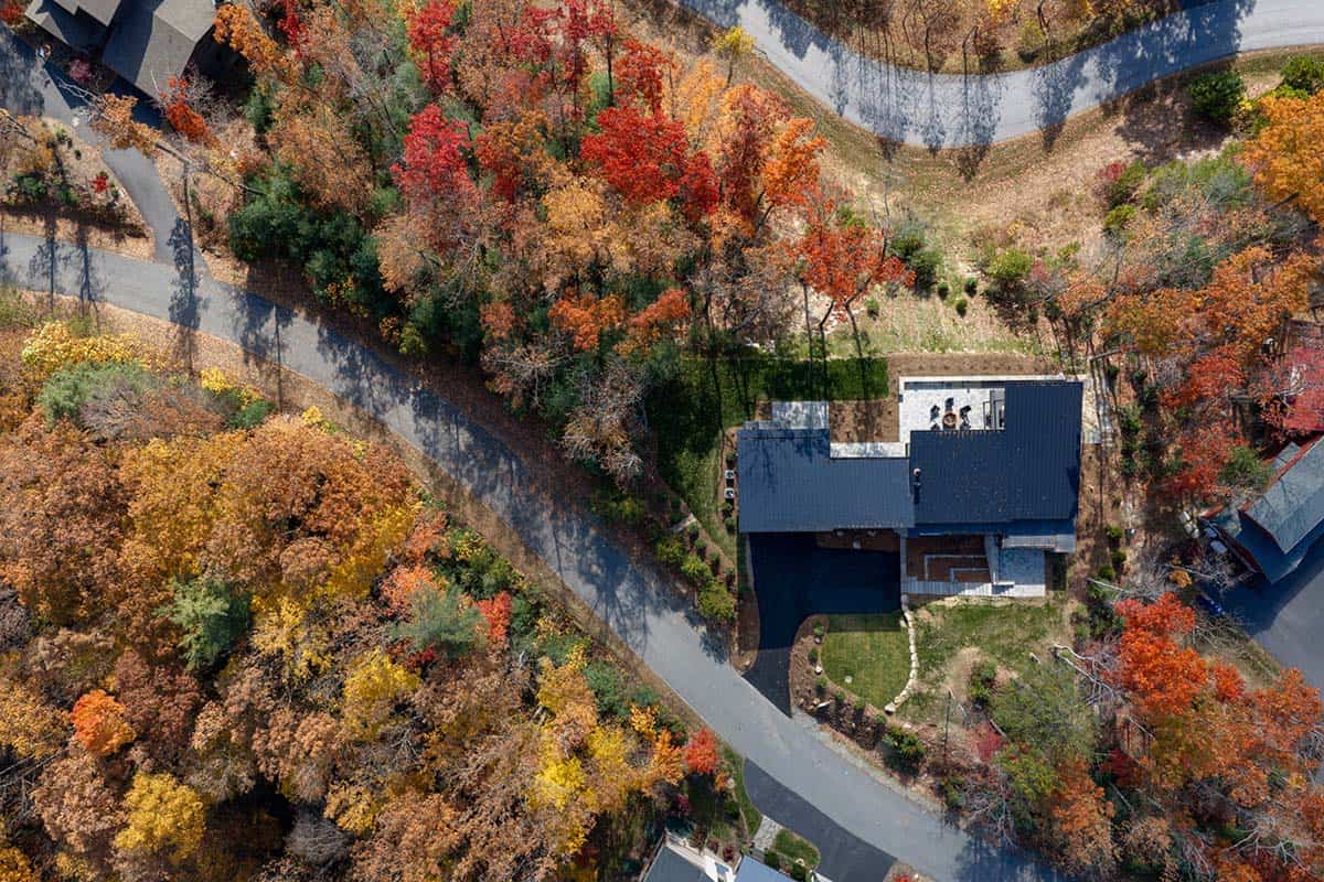 Aerial view of home's glass rear facade, deck, and stone patio surrounded by autumn foliage