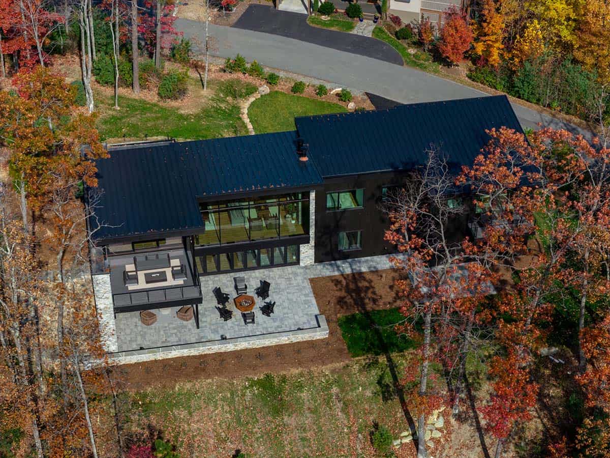 Aerial view of dark metal-roofed home with glass wall and patio amid fall trees