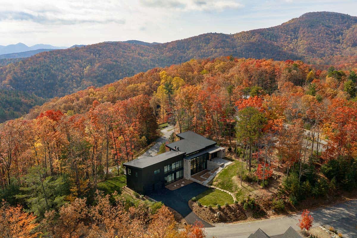 Aerial view of modern black home perched on forested mountainside with layered ridgelines beyond