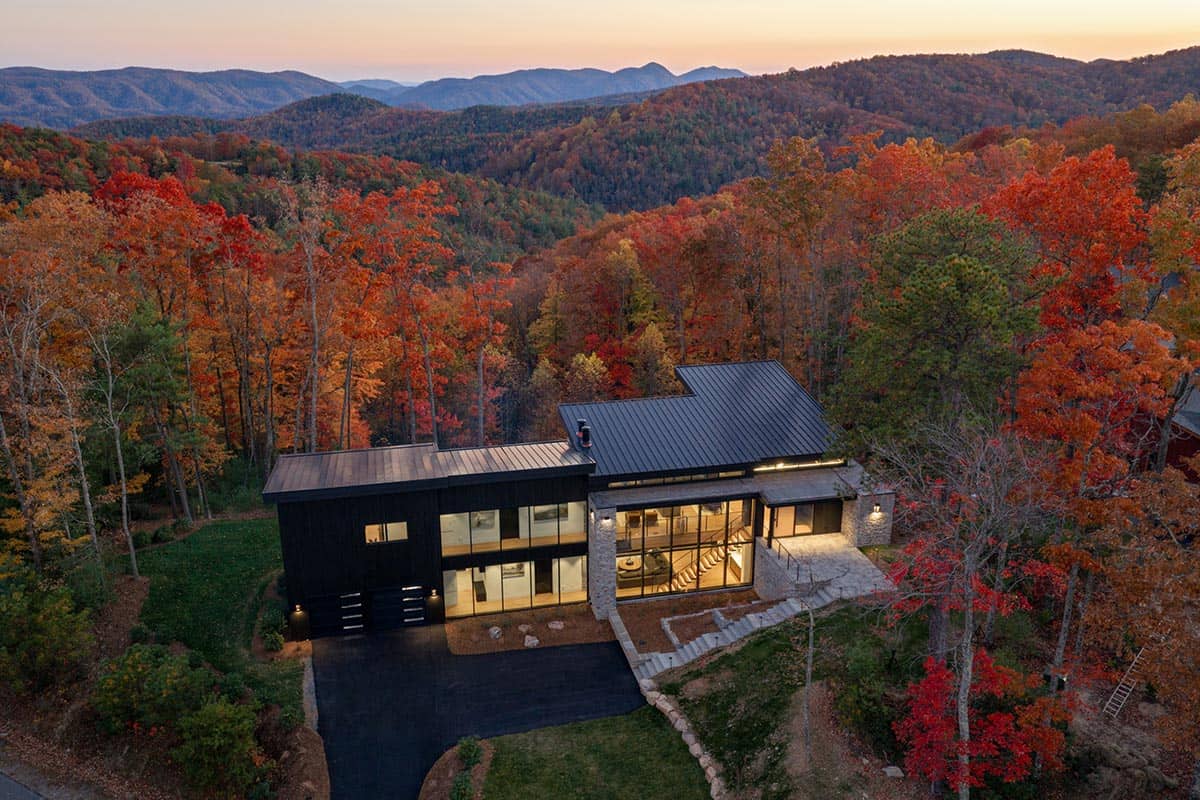 Aerial dusk view of dark modern home with lit interior amid red and orange foliage