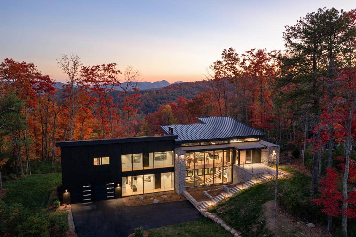 Aerial twilight view of illuminated home nestled in forest with mountain ridge silhouette