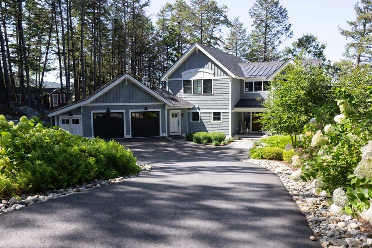 Driveway curves toward gray shingle cottage with hydrangeas and river stone edging