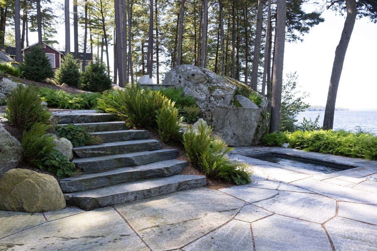 Stone patio with granite steps, large boulder, and spa beside the lake