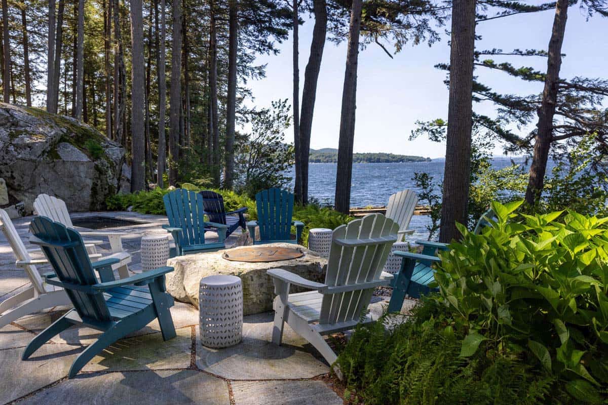 Adirondack chairs circle a boulder fire pit overlooking the lake