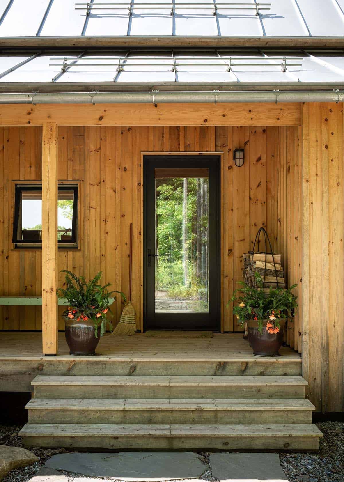 Cabin porch with black-framed glass door, potted ferns, and stacked firewood