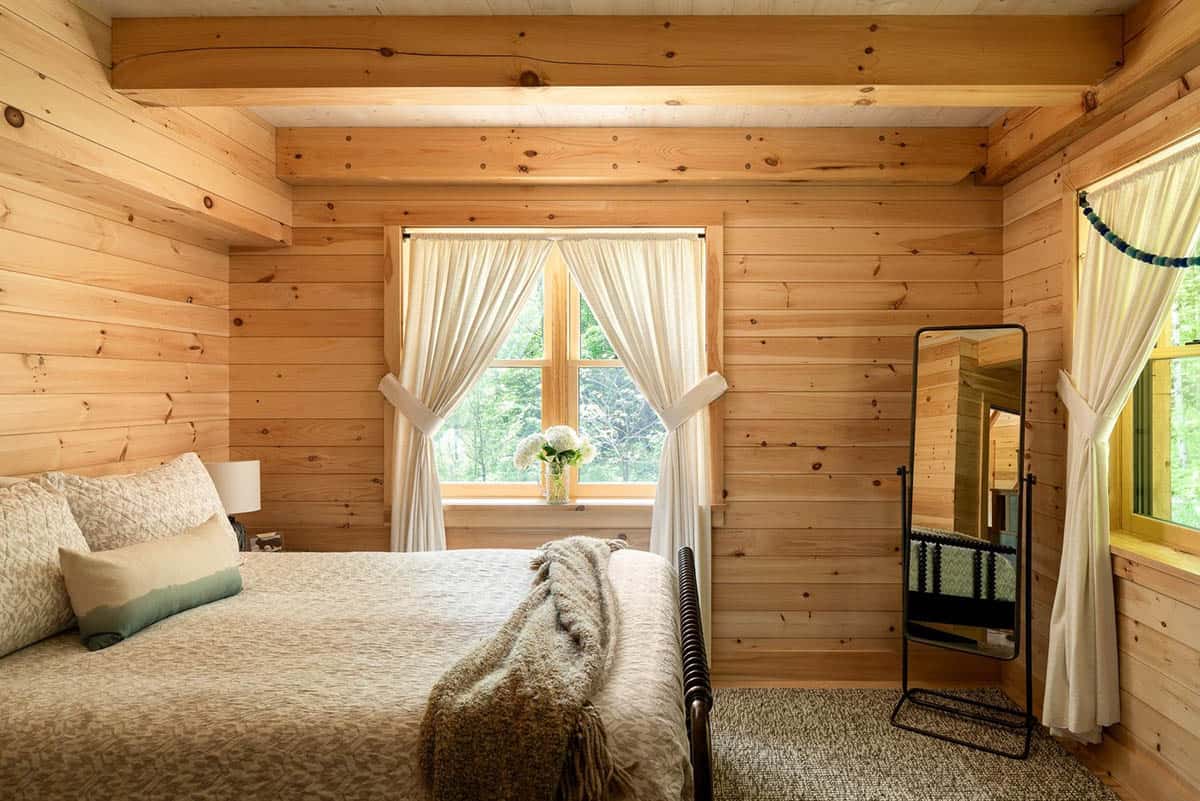 Wood-paneled bedroom with exposed beams, white curtains, and forest views