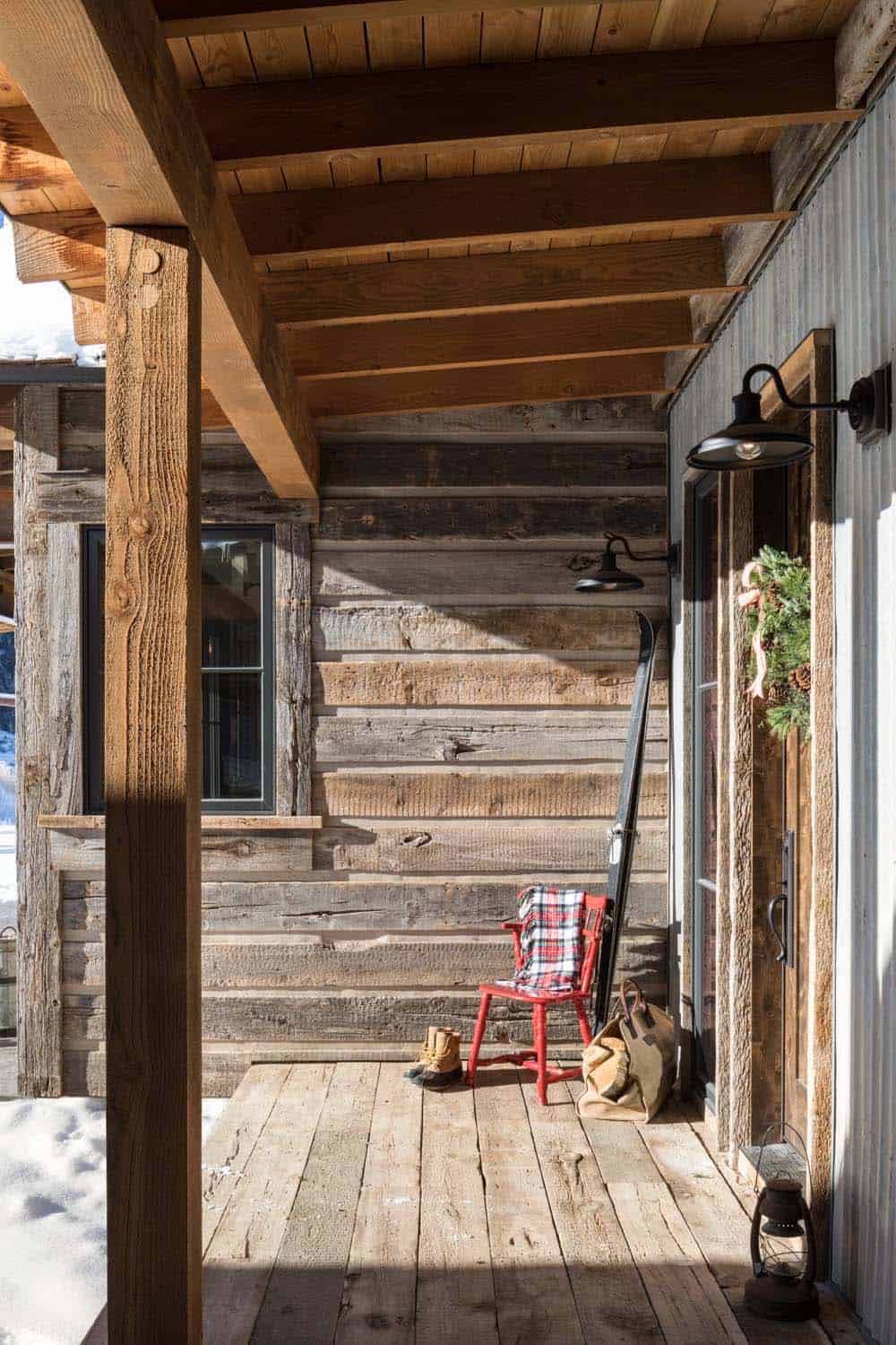 Covered wooden porch with red rocking chair, plaid blanket, skis, and wreath on barnwood siding