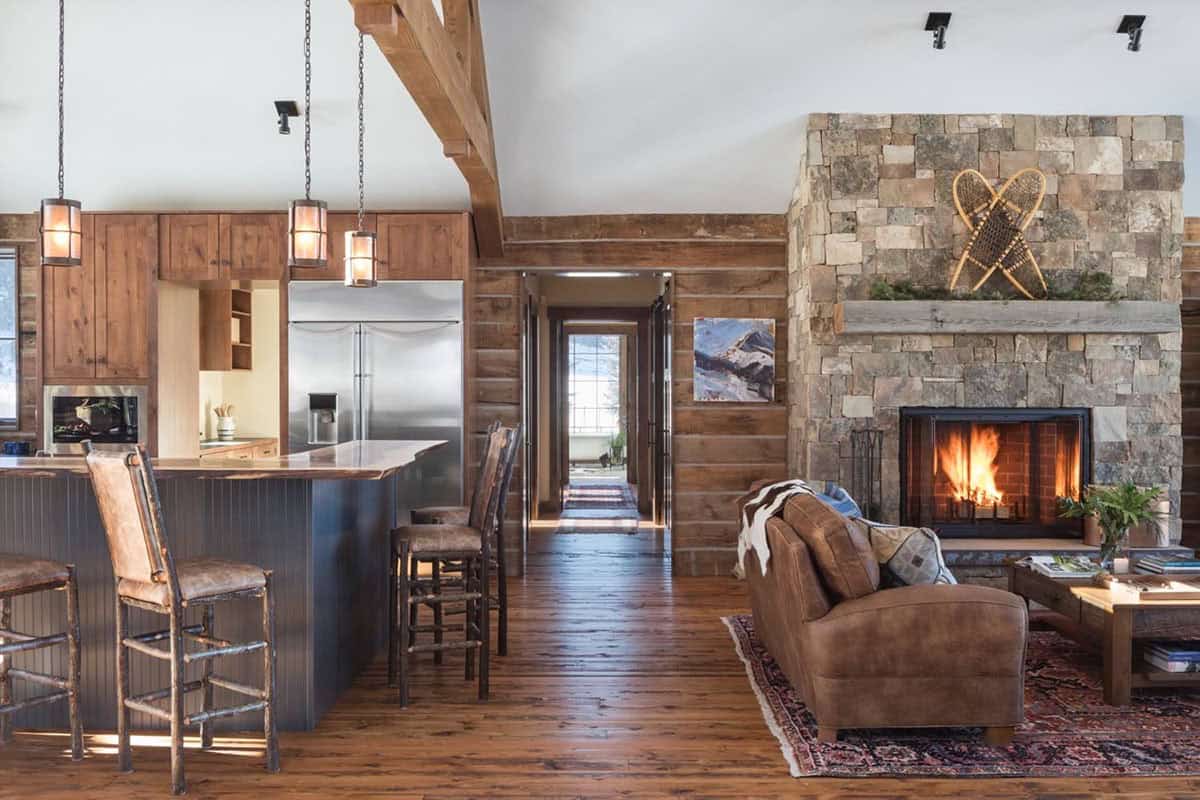 Open-concept living area with kitchen island, wood beams, and stone fireplace with fire lit