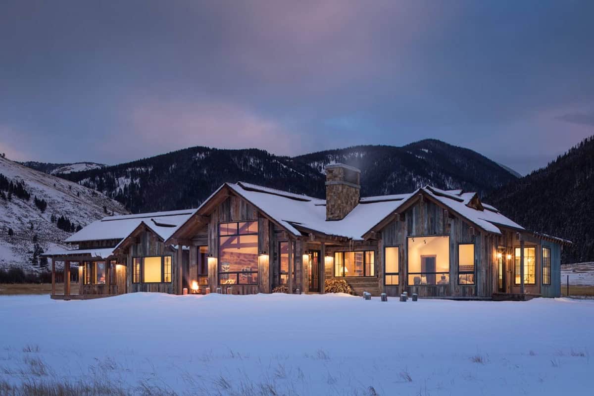 Rustic wood home exterior glowing with interior lights at dusk against snowy mountains