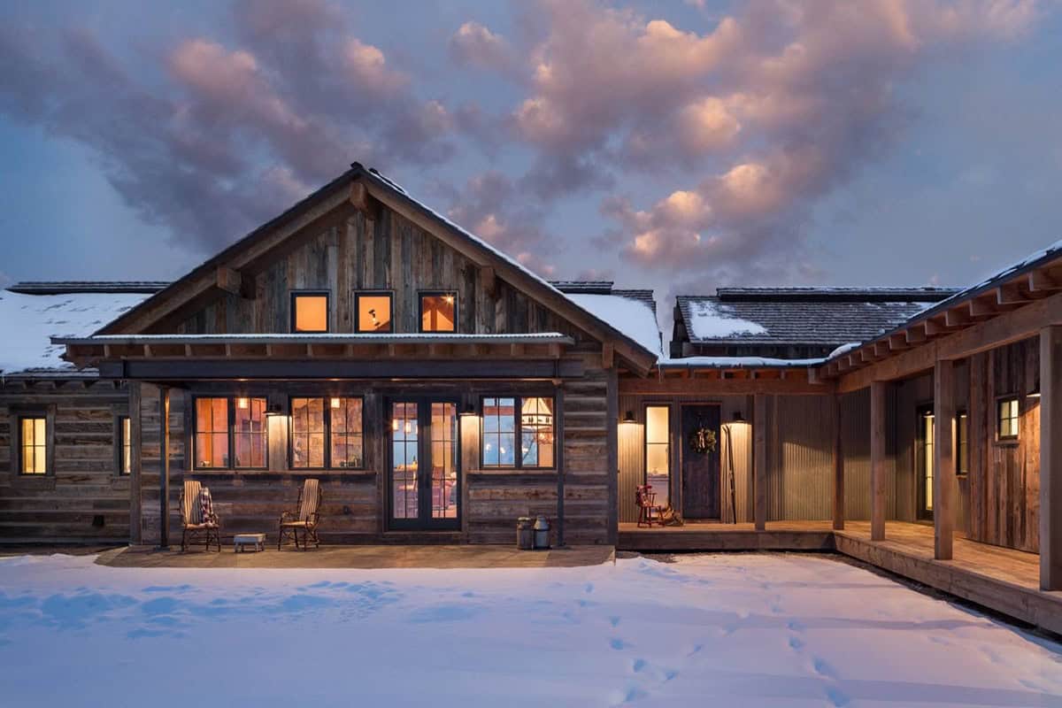 Twilight view of cabin exterior with lit windows, covered porch, and snow-covered ground