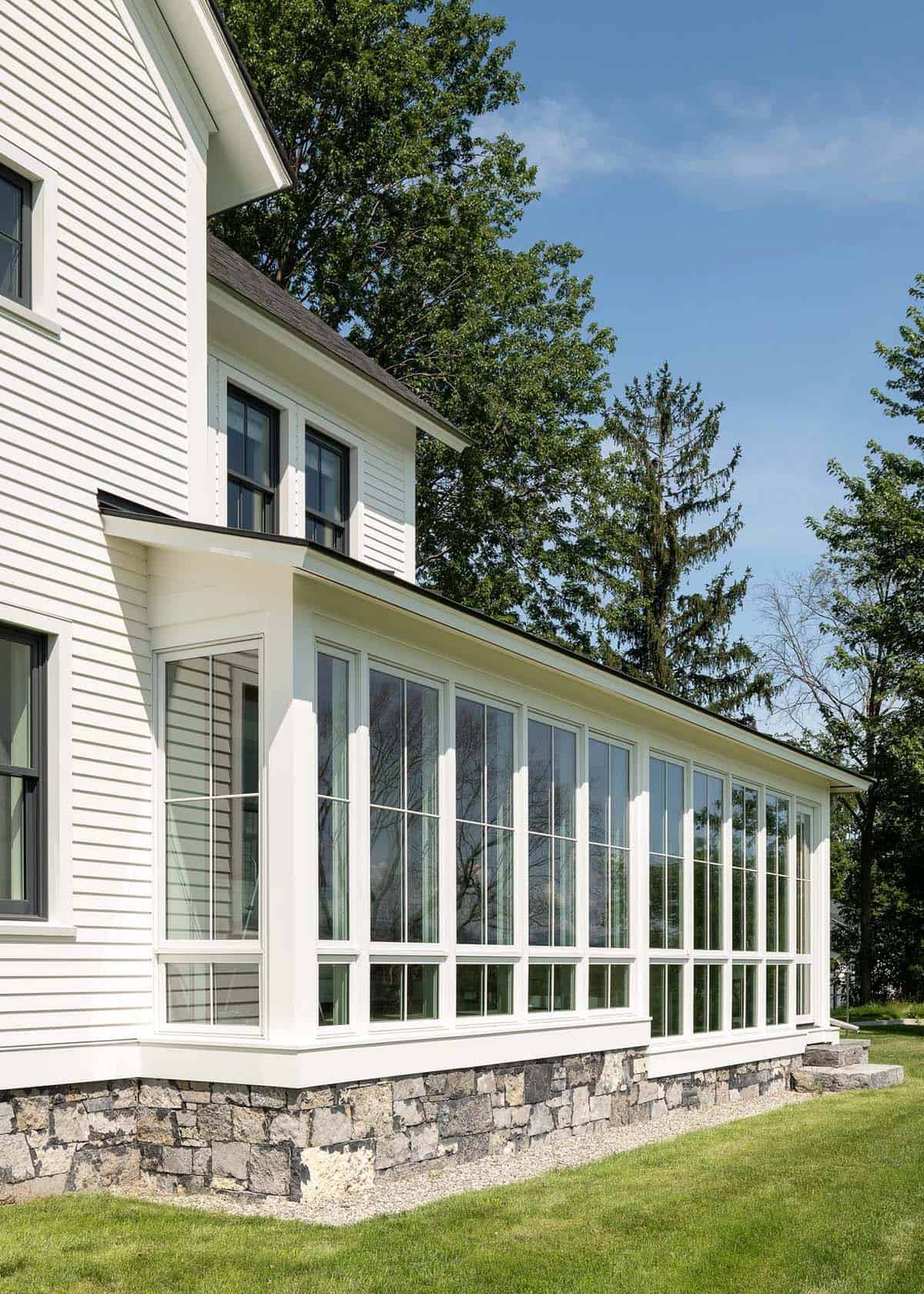 Glass-walled sunroom with white-trimmed windows on a fieldstone foundation