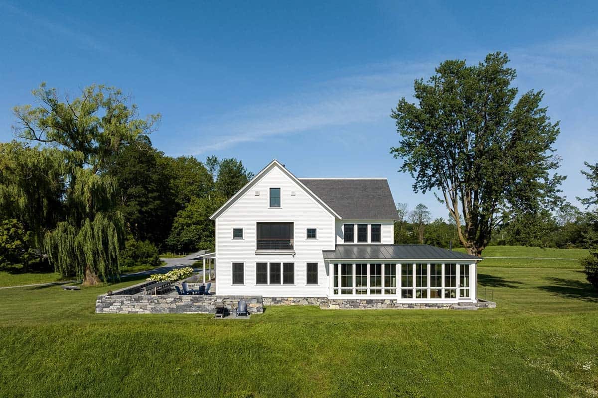Back elevation of the white farmhouse with glass sunroom and stone patio among mature trees