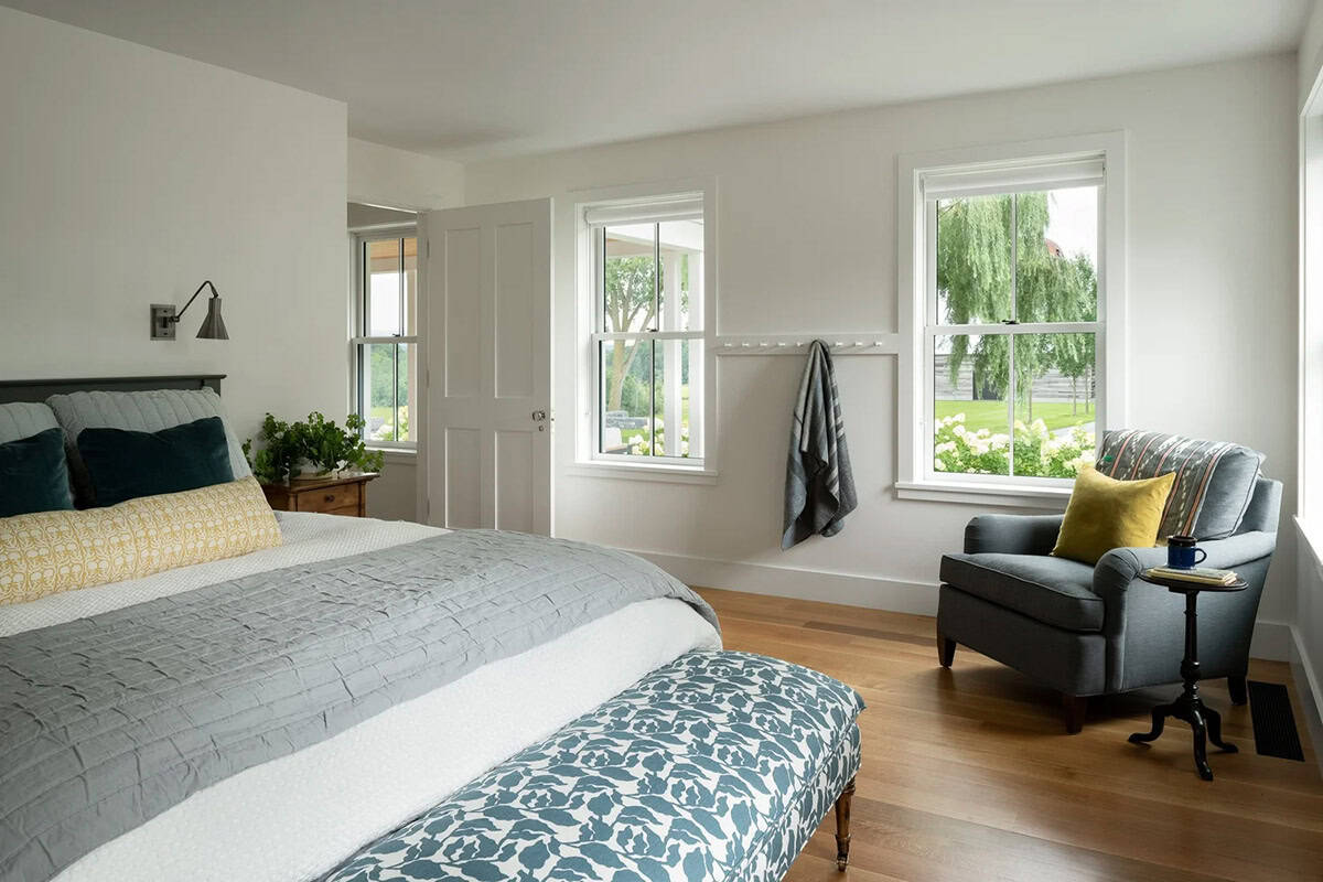 Guest bedroom with gray bedding, upholstered bench, and armchair beside windows facing hydrangeas