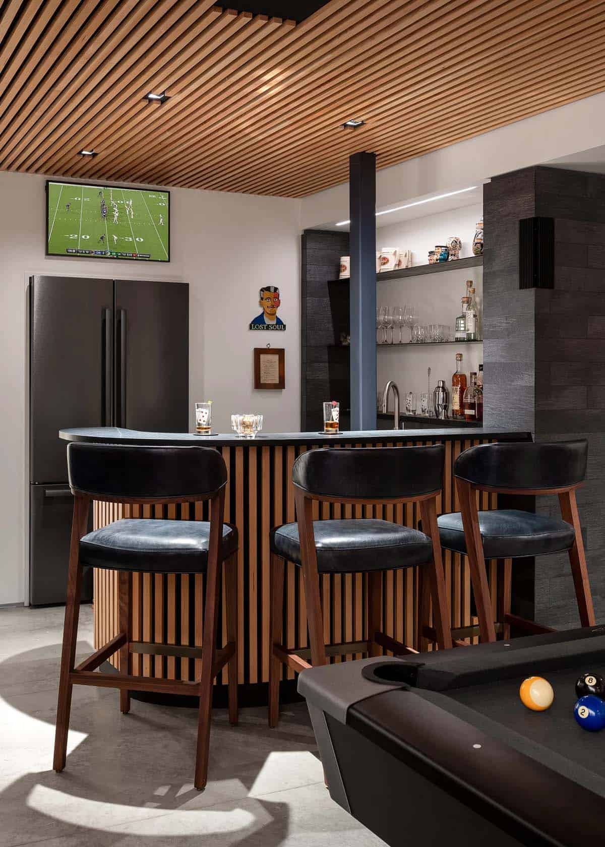 Basement bar with curved slatted wood front, leather stools, and open shelving