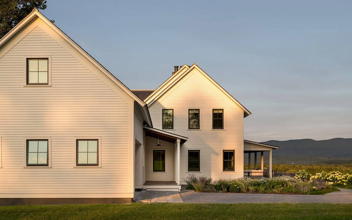 White clapboard farmhouse at sunset with black-framed windows, covered porch, and mountain backdrop