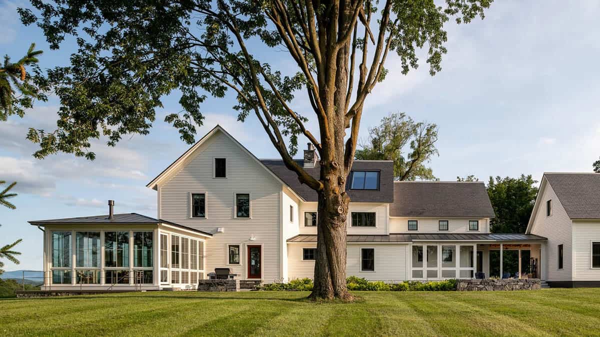 Back of the farmhouse behind a large tree with glass sunroom, red door, and covered porch