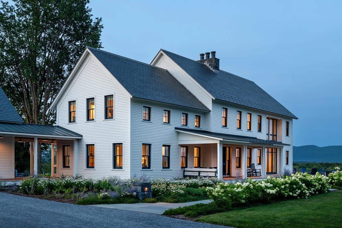 Farmhouse at dusk with glowing windows, covered porch, and blooming hydrangeas