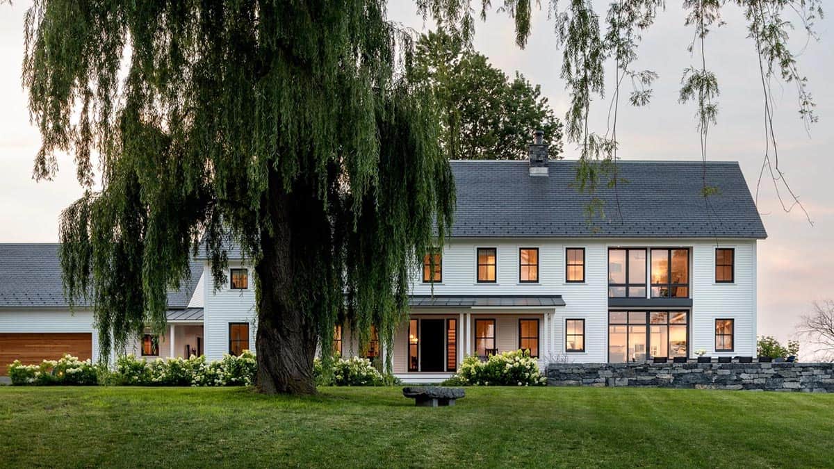 Front of the farmhouse at dusk behind a weeping willow with lit windows and stone wall
