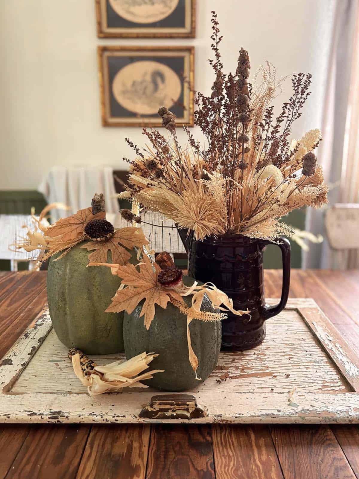 Dark pitcher with dried flowers beside hand-painted textured pumpkins on wood table