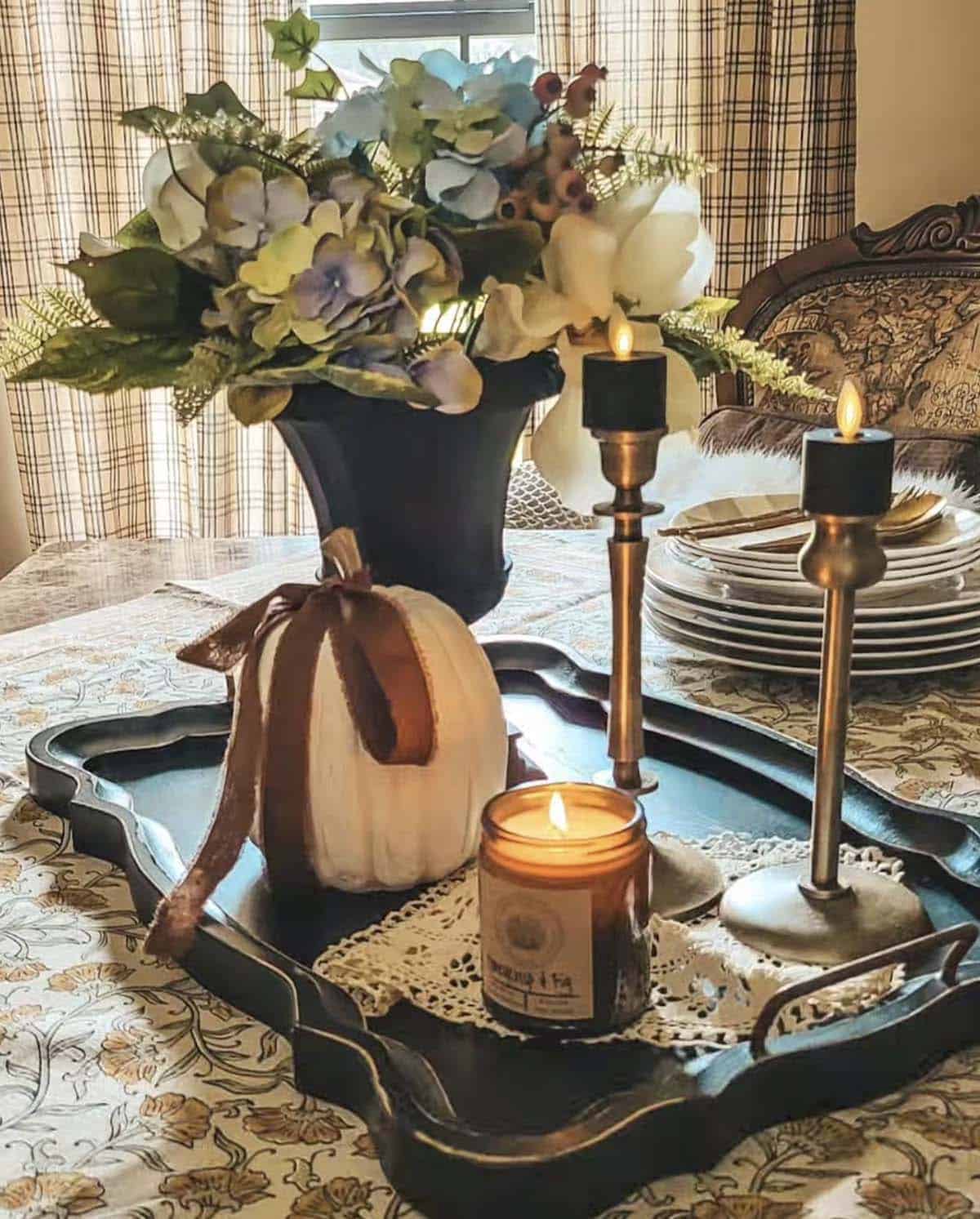 Black vase with florals beside velvet-ribbon pumpkin and candles on scalloped tray