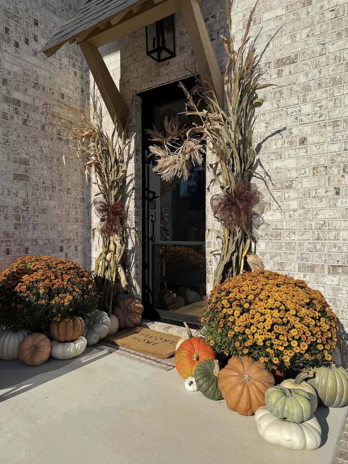 Fall entryway with black door, corn stalks, orange mums, and pumpkins