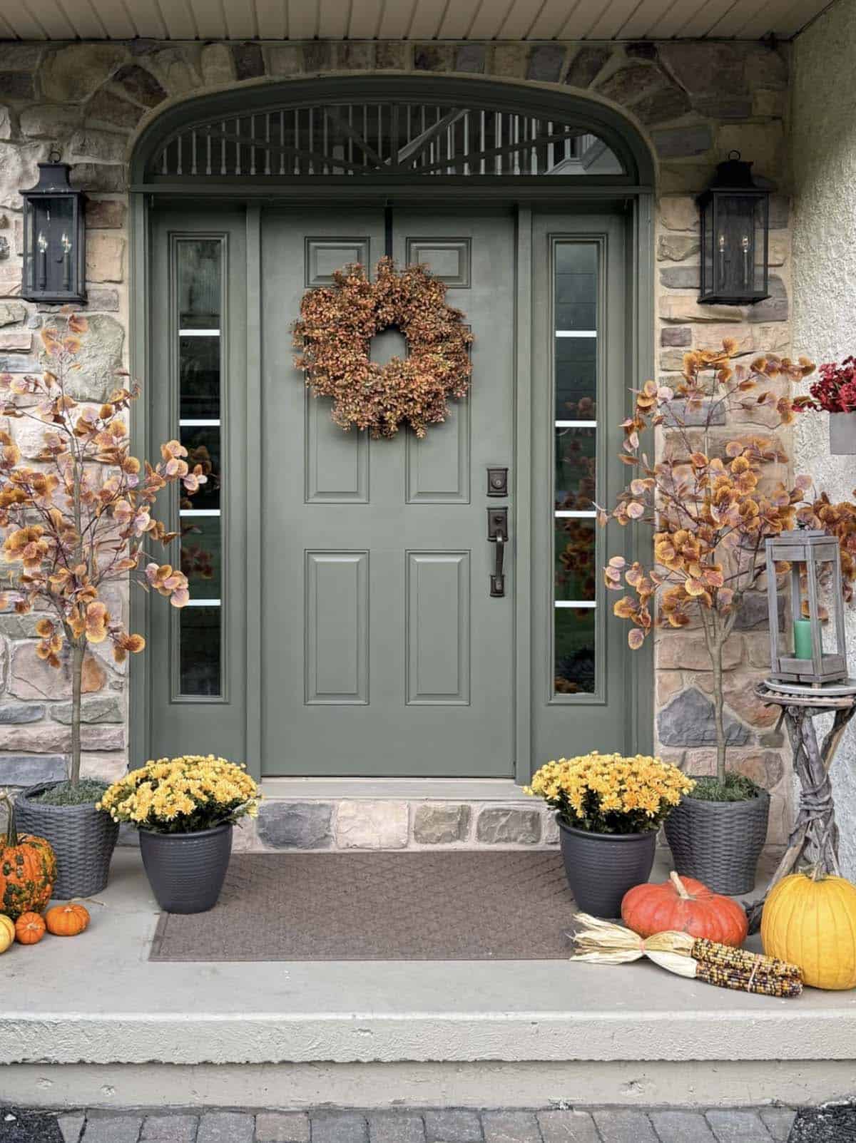 Green front door with autumn wreath, fall trees, yellow mums, and pumpkins