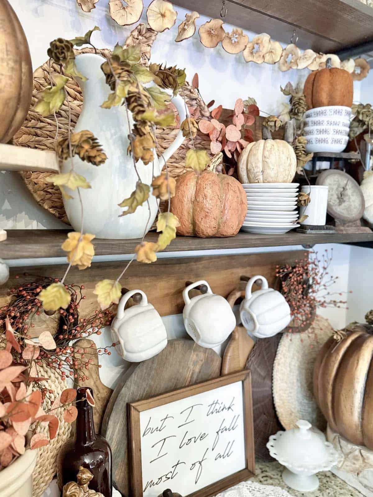 White pitcher with faux pumpkins, dried apple garland, and stacked plates on shelf