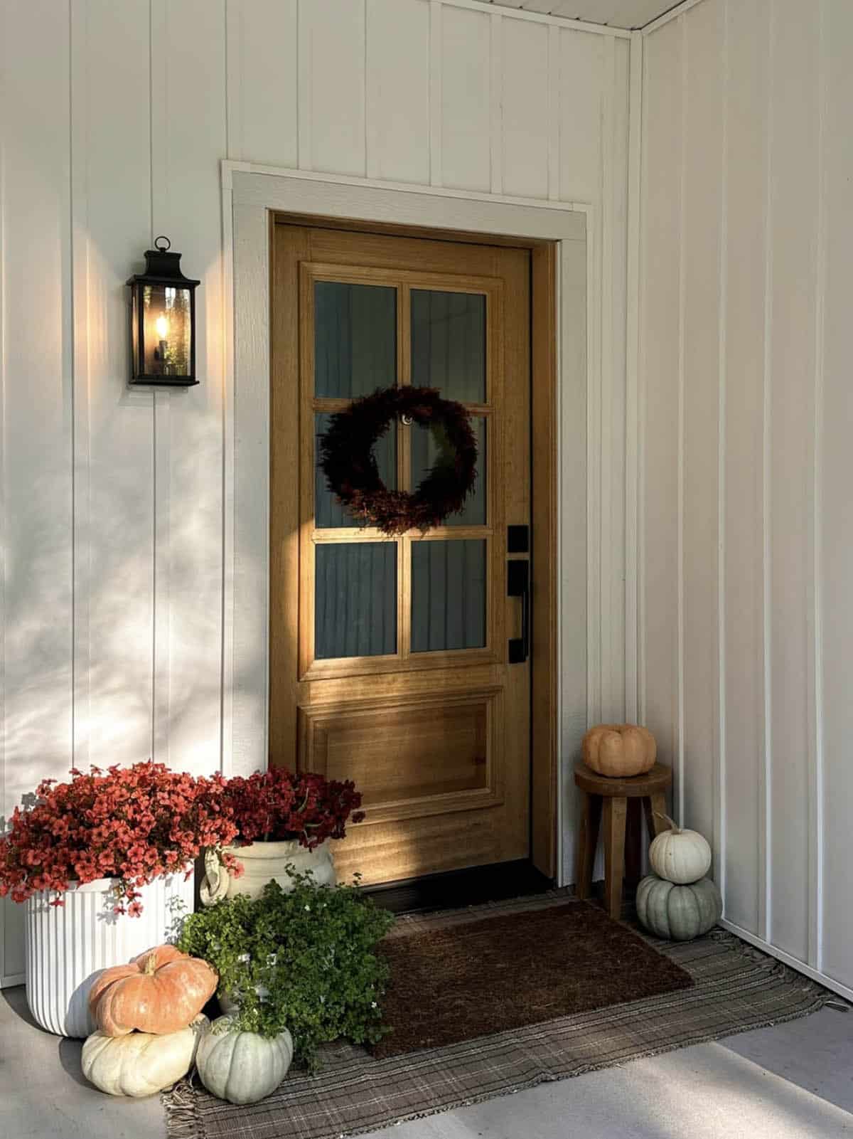 Wood front door with wreath, red flowers in white planters, and pumpkins