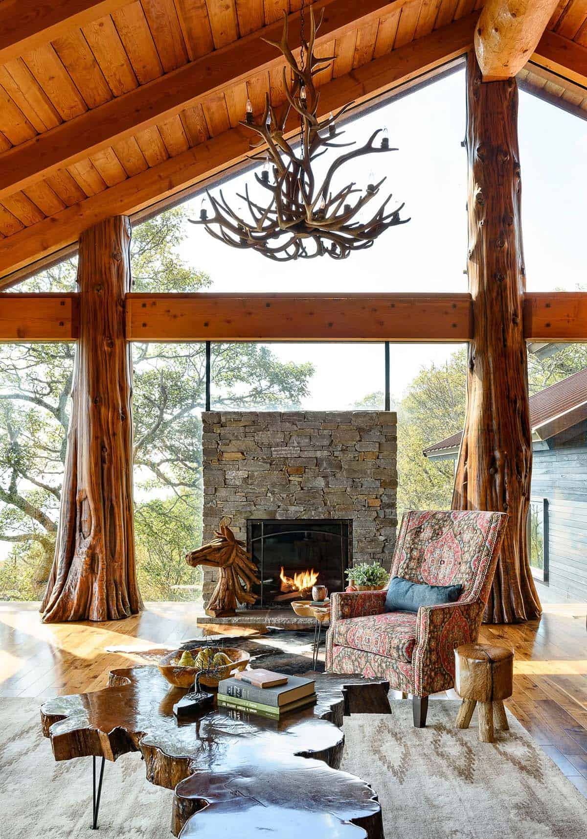 Antler chandelier hangs above a stone fireplace and tree trunk columns