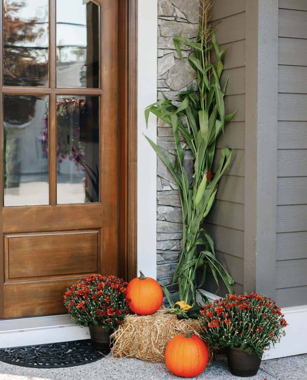 Porch corner with hay bale, red mums, pumpkins, and green corn stalk