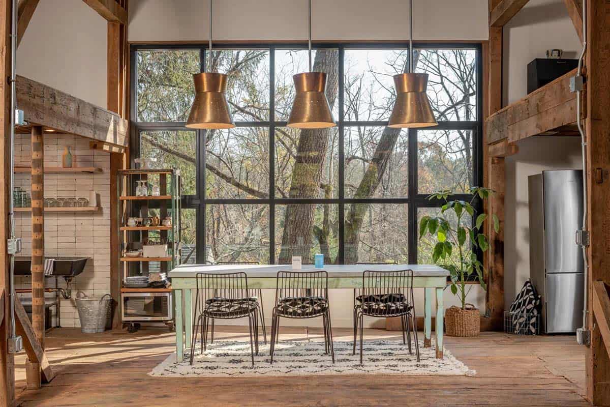 Open-concept dining area with a long table, copper pendant lights, and floor-to-ceiling windows overlooking trees