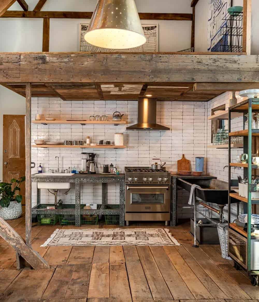 Rustic kitchen under a wood loft, featuring white tile backsplash, industrial shelving, and stainless range