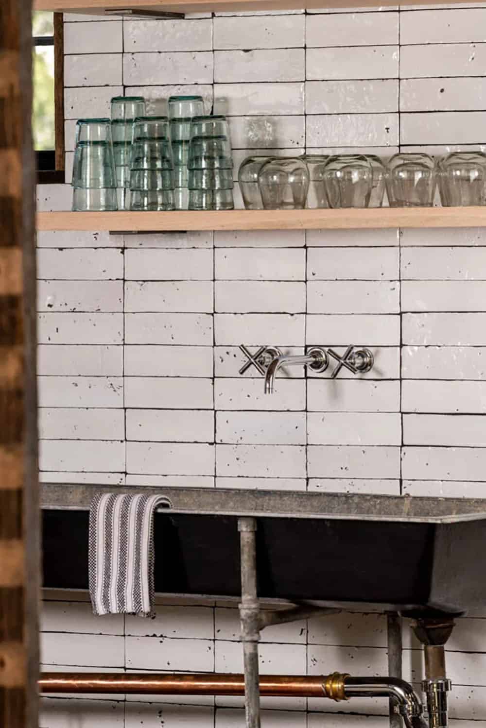 Close-up of whitewashed brick tile backsplash with wall-mounted faucet and vintage glassware on open shelves