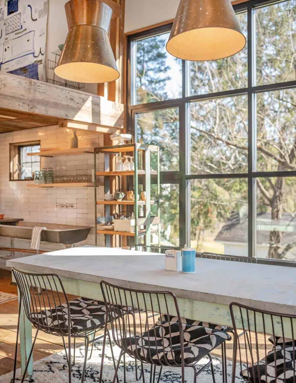 Dining nook with mismatched wire chairs, concrete-top table, and large barn-style windows letting in natural light