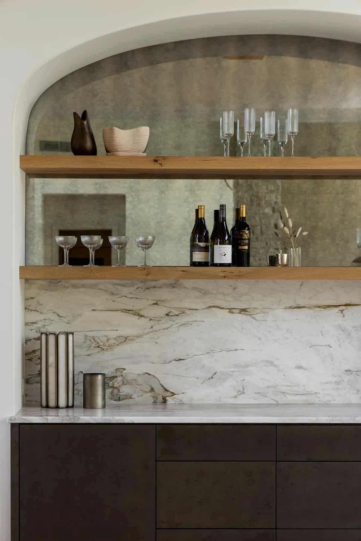 Mirrored arched shelves with wine bottles and coupe glasses above marble backsplash