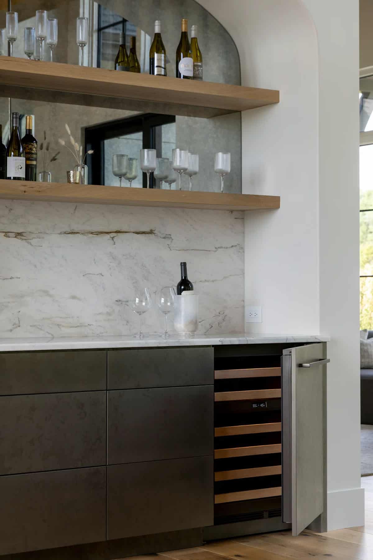 Built-in wine refrigerator below marble backsplash and mirrored open shelves