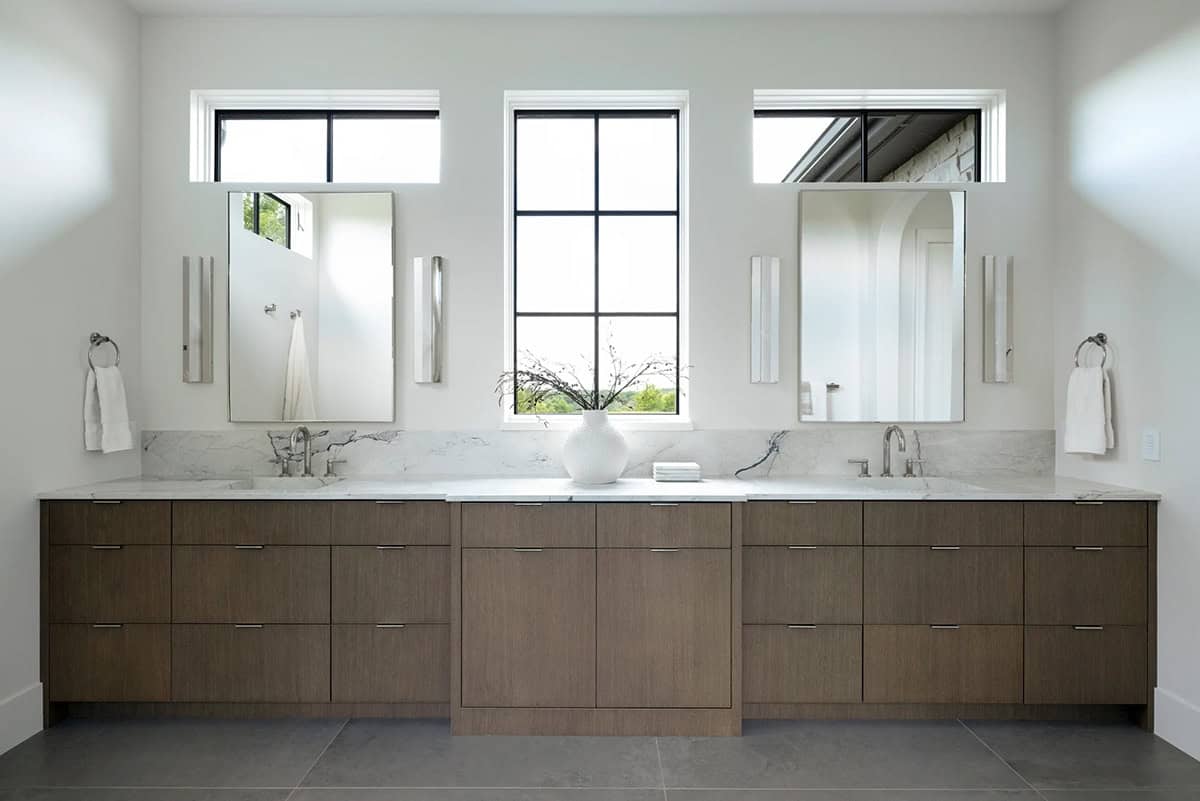 Double vanity with walnut-toned cabinetry, marble counters, and three windows