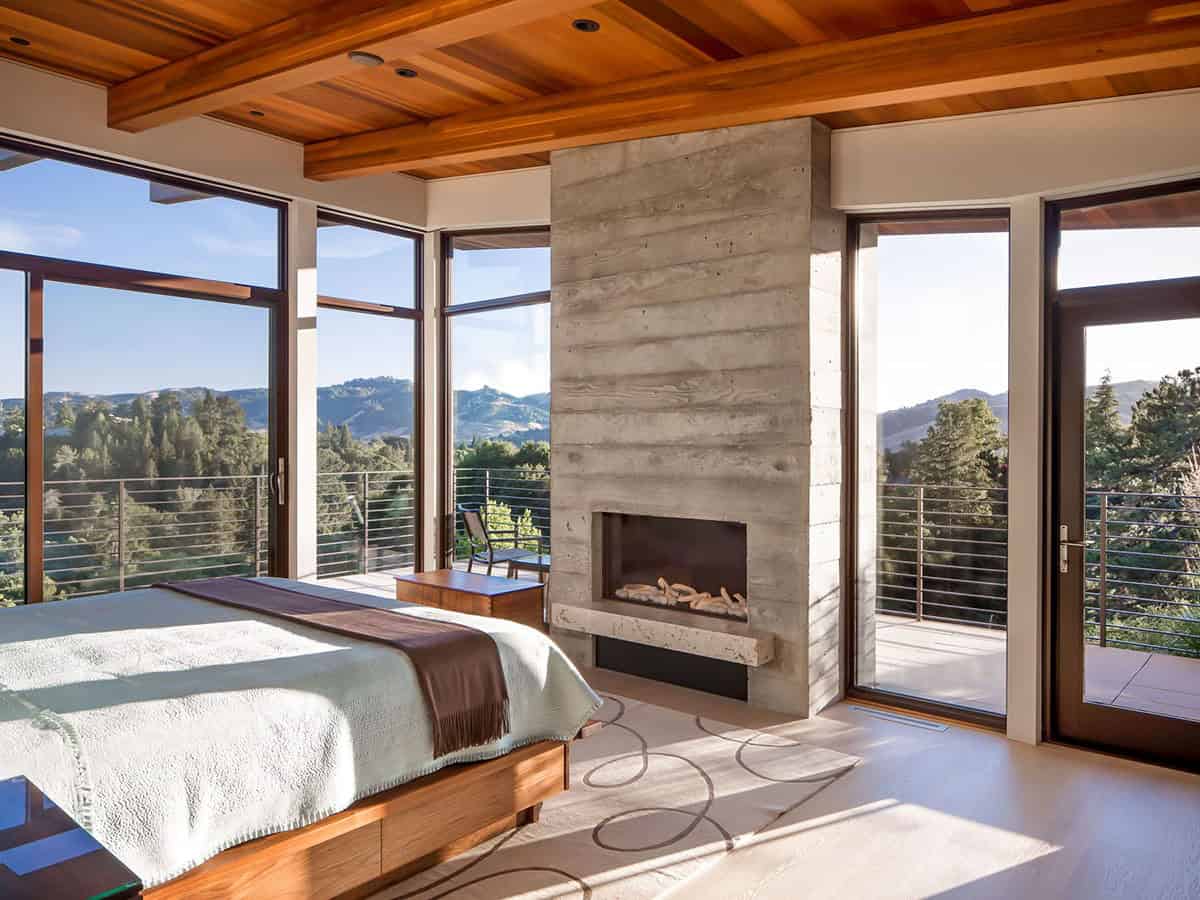 Owner's bedroom with corner windows framing mountain views beside concrete fireplace