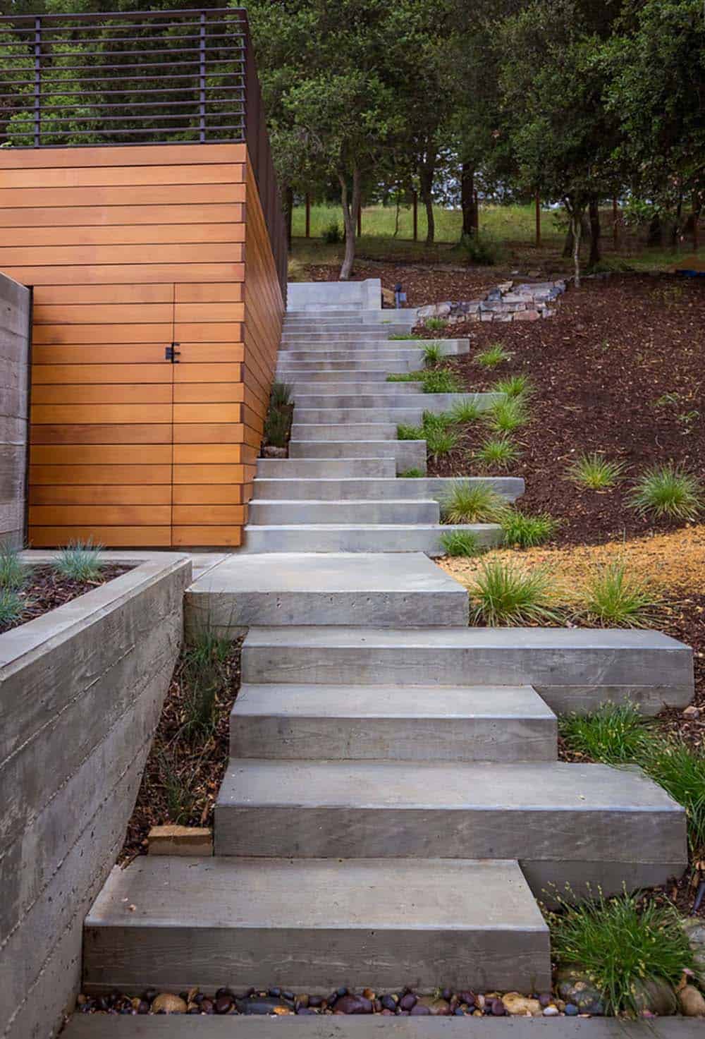 Concrete steps climb wooded hillside past ornamental grasses toward upper yard