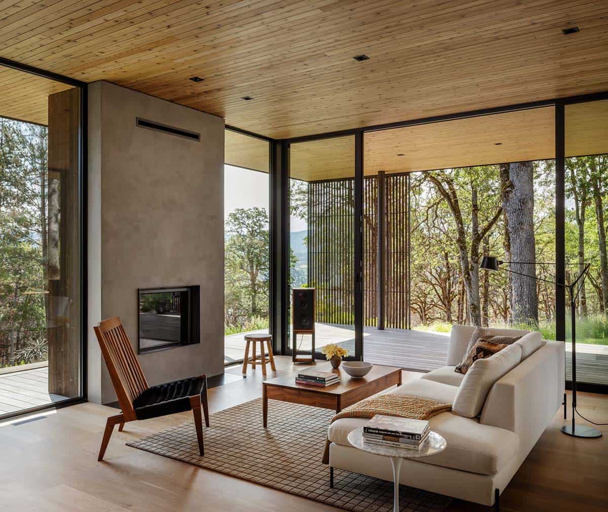 Living room seating with a fireplace and oak forest views through glass 