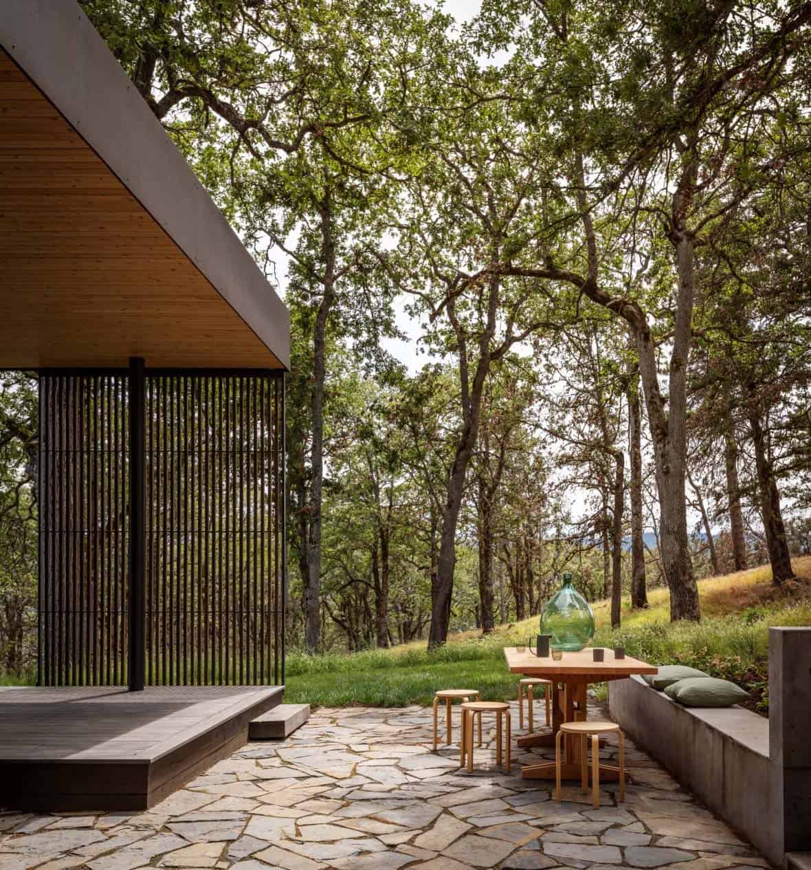 Stone patio with wood dining table and concrete bench beneath oak trees