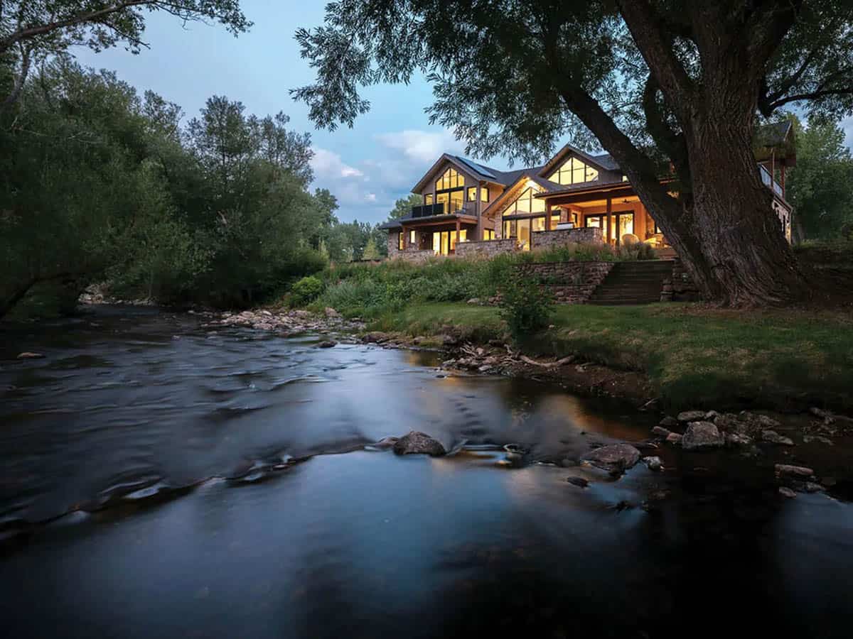 Glowing rear of the home at dusk beside a rocky creek
