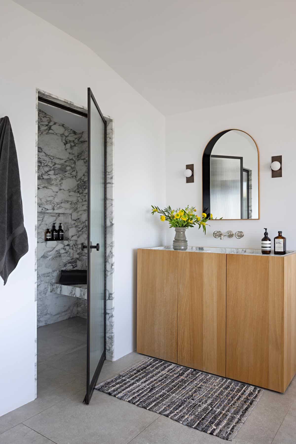 Bathroom with oak vanity, arched mirror, and marble shower with glass door