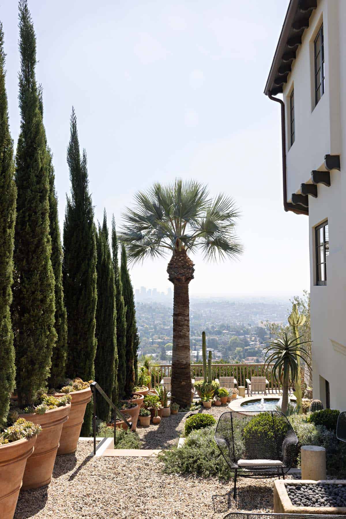 Hillside garden with potted cypress trees, palm tree, and city views