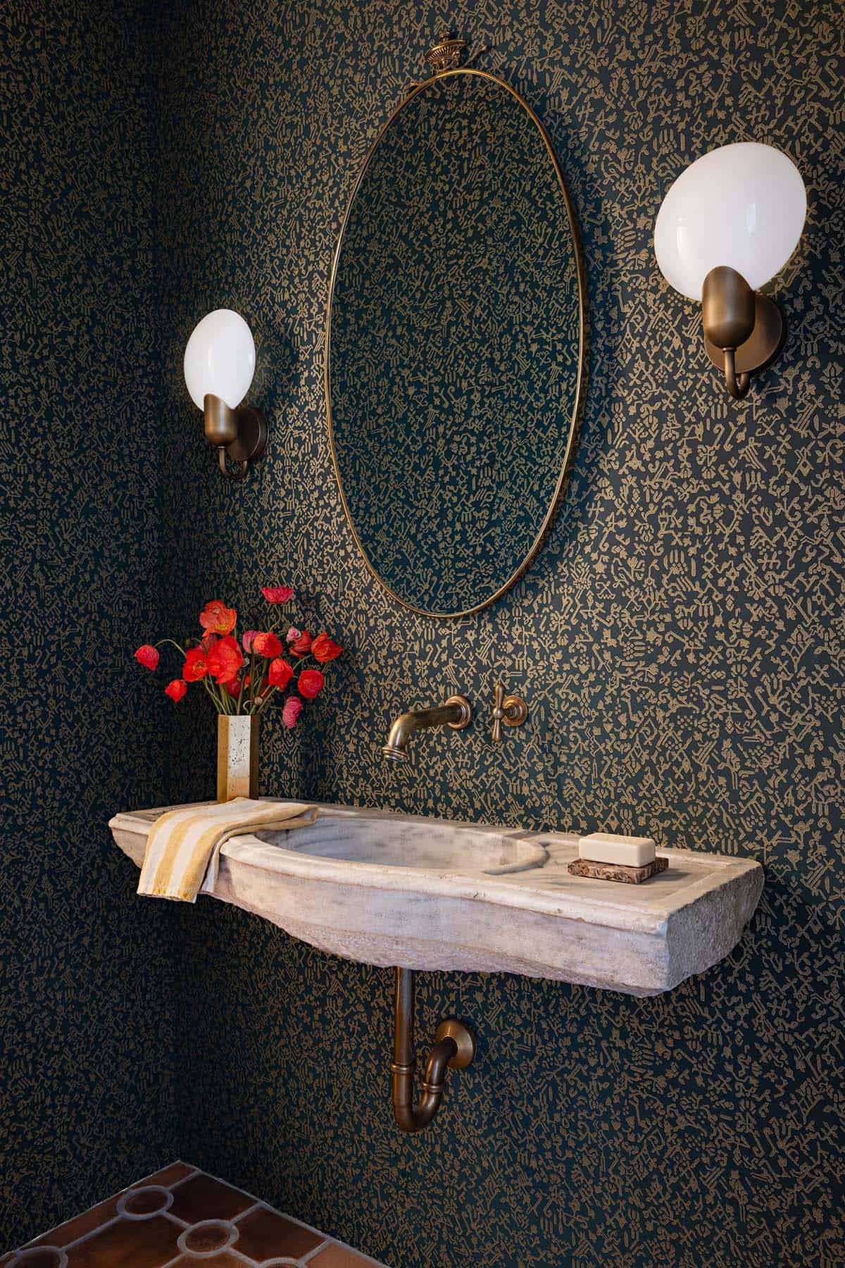 Powder room with antique stone sink, oval brass mirror, and patterned wallpaper